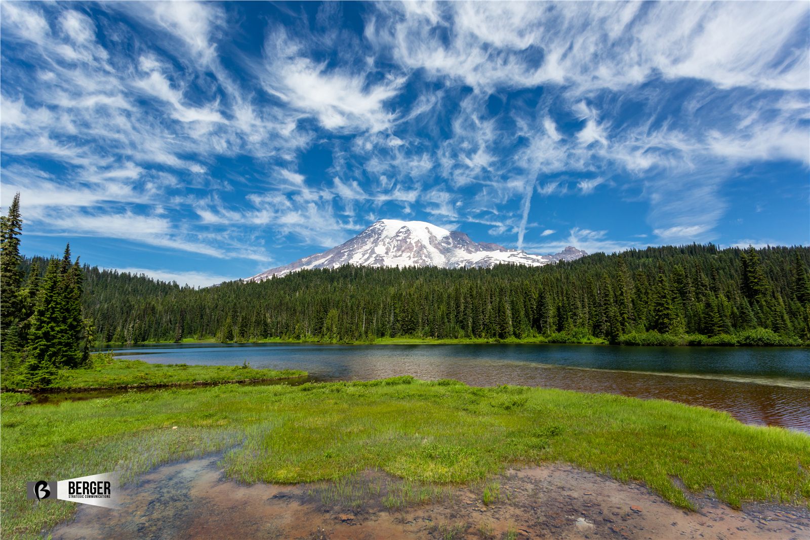Mount Rainier above a green meadow and mountain lake beneath textured clouds